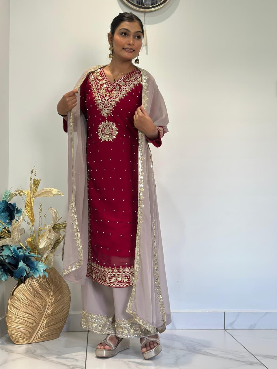 Woman wearing a red and gold embroidered traditional outfit with a white dupatta, standing indoors. Red Palazzo Suit Set Roohri