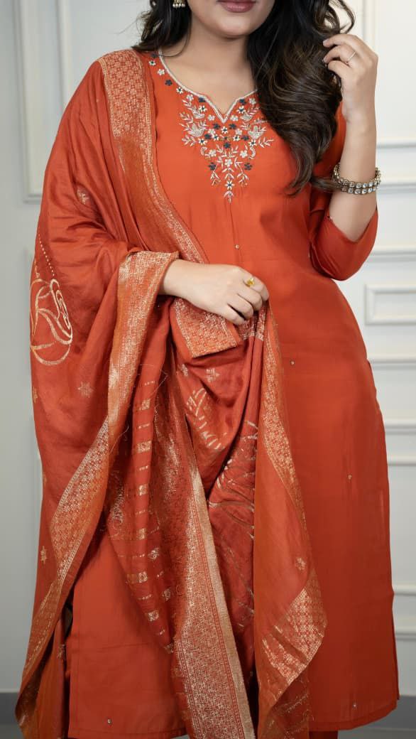 Woman wearing an orange traditional outfit with embroidery, holding a matching dupatta. Orange hand embroidered suit set Roohri