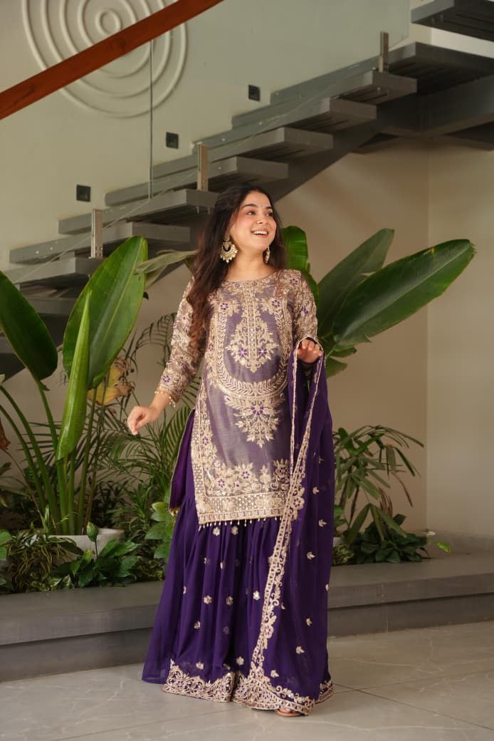 Woman in a traditional outfit standing indoors with plants and a staircase in the background. Purple palazzo suit set Roohri