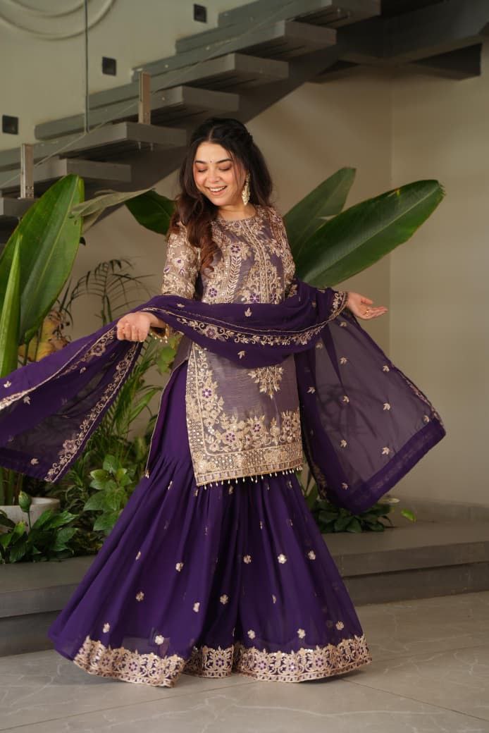 Woman in a traditional purple and white outfit with a matching dupatta, standing indoors with plants in the background. Purple palazzo suit set Roohri