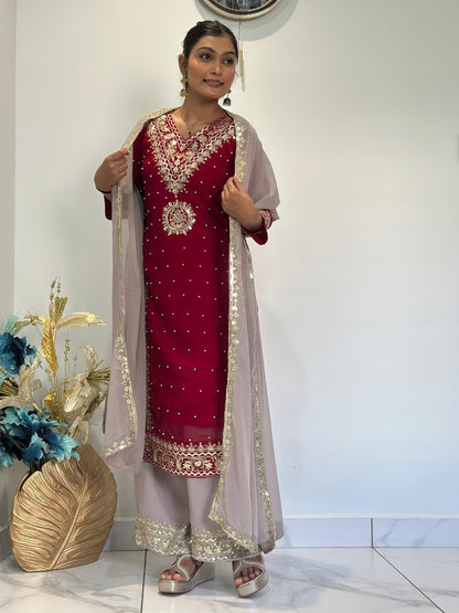 Woman wearing a red and gold embroidered traditional outfit with a white dupatta, standing indoors. Red Palazzo Suit Set Roohri