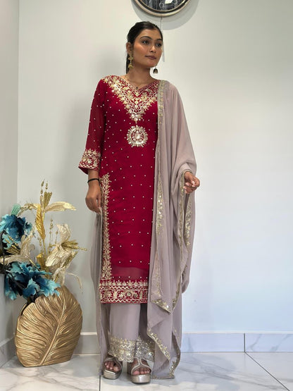 Woman wearing a red and gold traditional outfit with a beige dupatta, standing indoors. Red Palazzo Suit Set Roohri